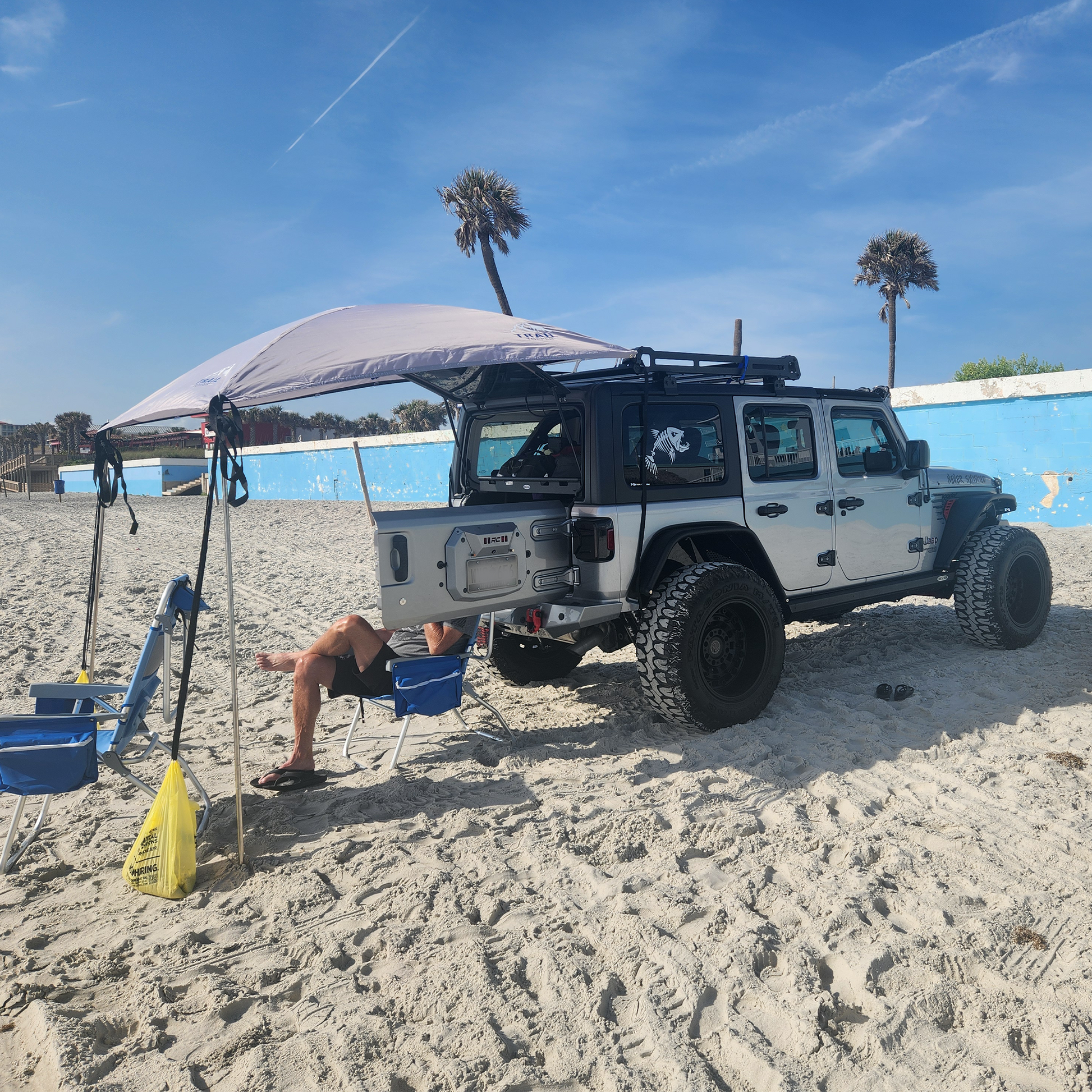 Jeep Wrangler  on a sandy beach with a person sitting under a JFTops Adventure Trail Topper shade awning