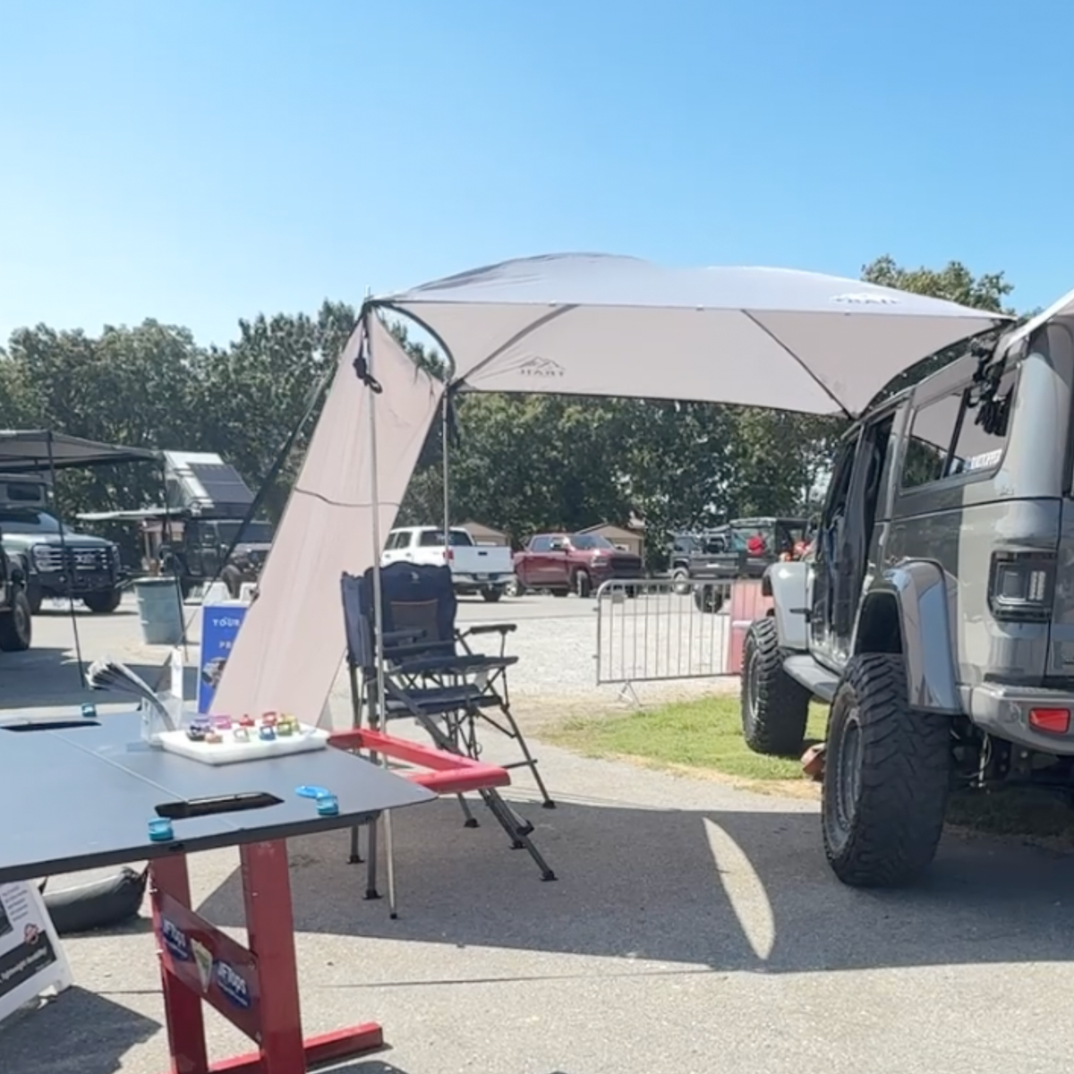 Outdoor setup with a trail topper awning, side walls, table, and chairs near a Jeep JT in an open area.