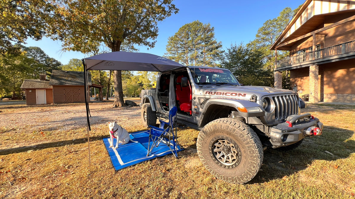 A silver Jeep Rubicon parked outdoors in a yard with an Adventure Trail Topper, blue folding chair, and blue mat on the grass. The vehicle has its doors removed, and a dog is sitting on the blue mat nearby.