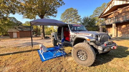 A silver Jeep Rubicon parked outdoors in a yard with an Adventure Trail Topper, blue folding chair, and blue mat on the grass. The vehicle has its doors removed, and a dog is sitting on the blue mat nearby.