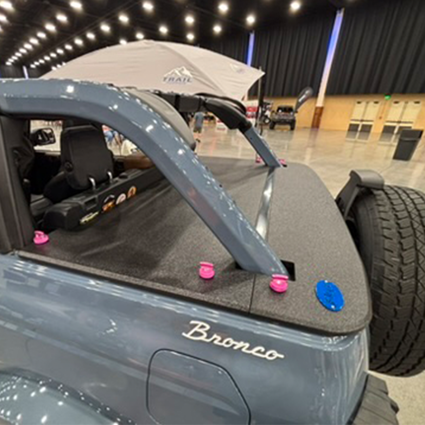 Cactus Gray Ford Bronco with pink Rockworkx Thumbscrews and JFTops rhino lined cargo cover on a showroom floor.