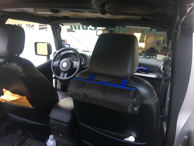 A black and blue trimmed dash poncho bag with blue straps hanging from the headrest of a black leather vehicle seat, shown inside a vehicle cabin.