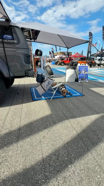 JT Gladiator with Overland Adventure for dog shade, folding chairs, and a dog lying on a blue mat on the asphalt under a partly cloudy sky.