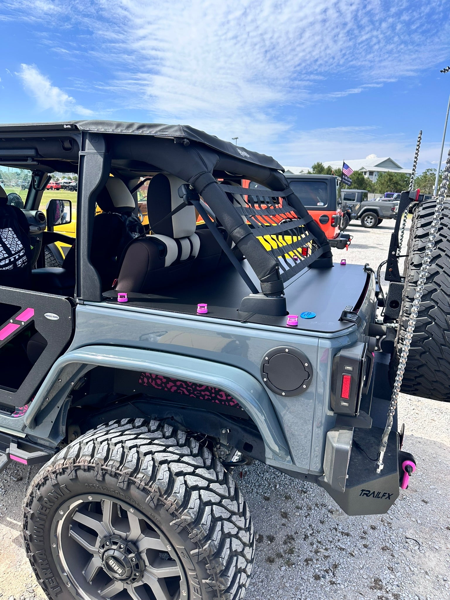 A gray off-road vehicle with a black soft top and roll bar, photographed outdoors at an event with JFtops 2025 aluminum softtop cargo cover. The vehicle has pink rockworkx thumbscrew accents, large off-road tires, and a black front bumper.