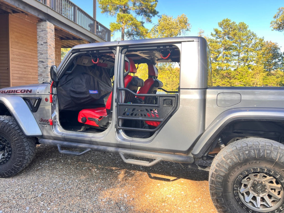 A silver Jeep Gladiator pickup truck with its doors and side cargo area removed, showing a red and black interior roll bar and seat covers installed. Dash Poncho protecting the dashboard. The truck is parked outdoors on a gravel surface next to a wooden cabin with trees in the background.
