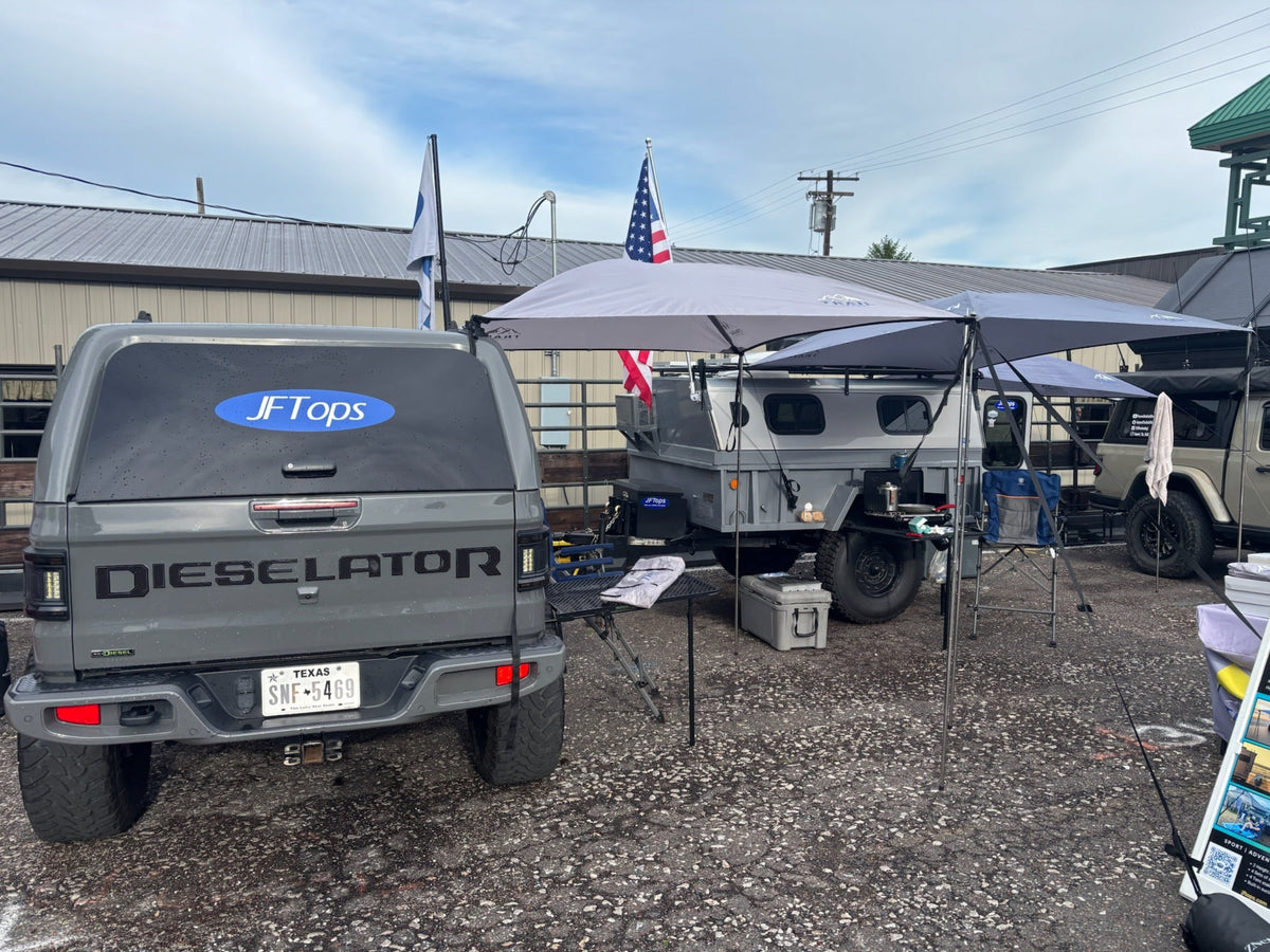 A JT Dieselator Jeep with a JFTops Trail Topper off the side for shade and rain protection, parked at an outdoor Moore Expo event next to a pop-up camper under another Trail Topper