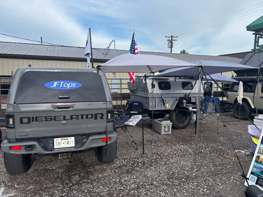 A JT Dieselator Jeep with a JFTops Trail Topper off the side for shade and rain protection, parked at an outdoor Moore Expo event next to a pop-up camper under another Trail Topper