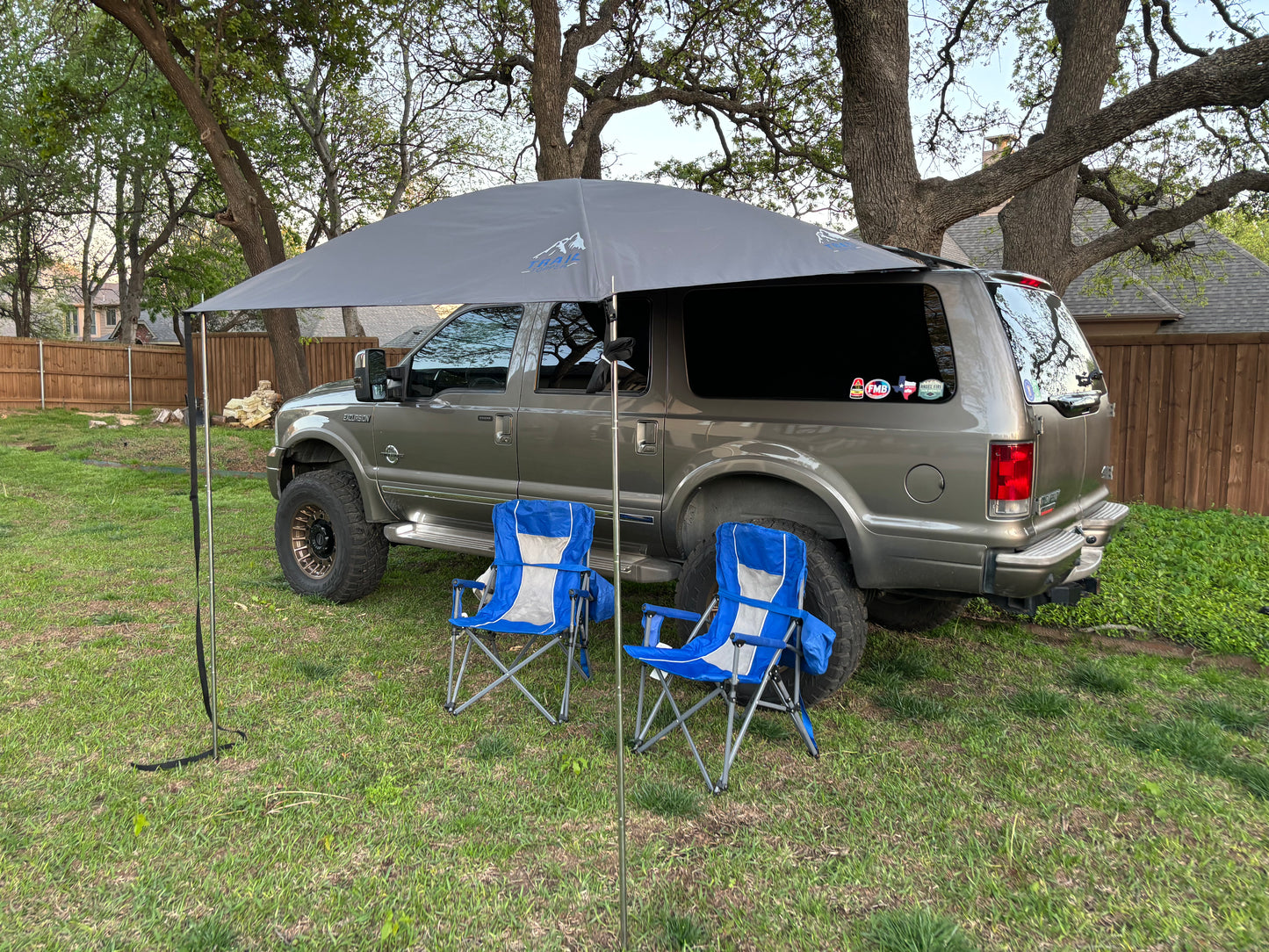 A shaded area with a JFTops 8x7 portable shade awning set up on a grassy field near a vehicle, with two chairs under the awning.