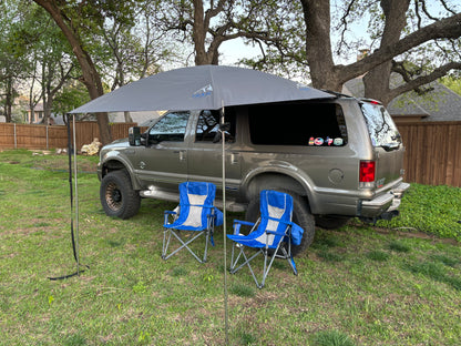 A shaded area with a JFTops 8x7 portable shade awning set up on a grassy field near a vehicle, with two chairs under the awning.