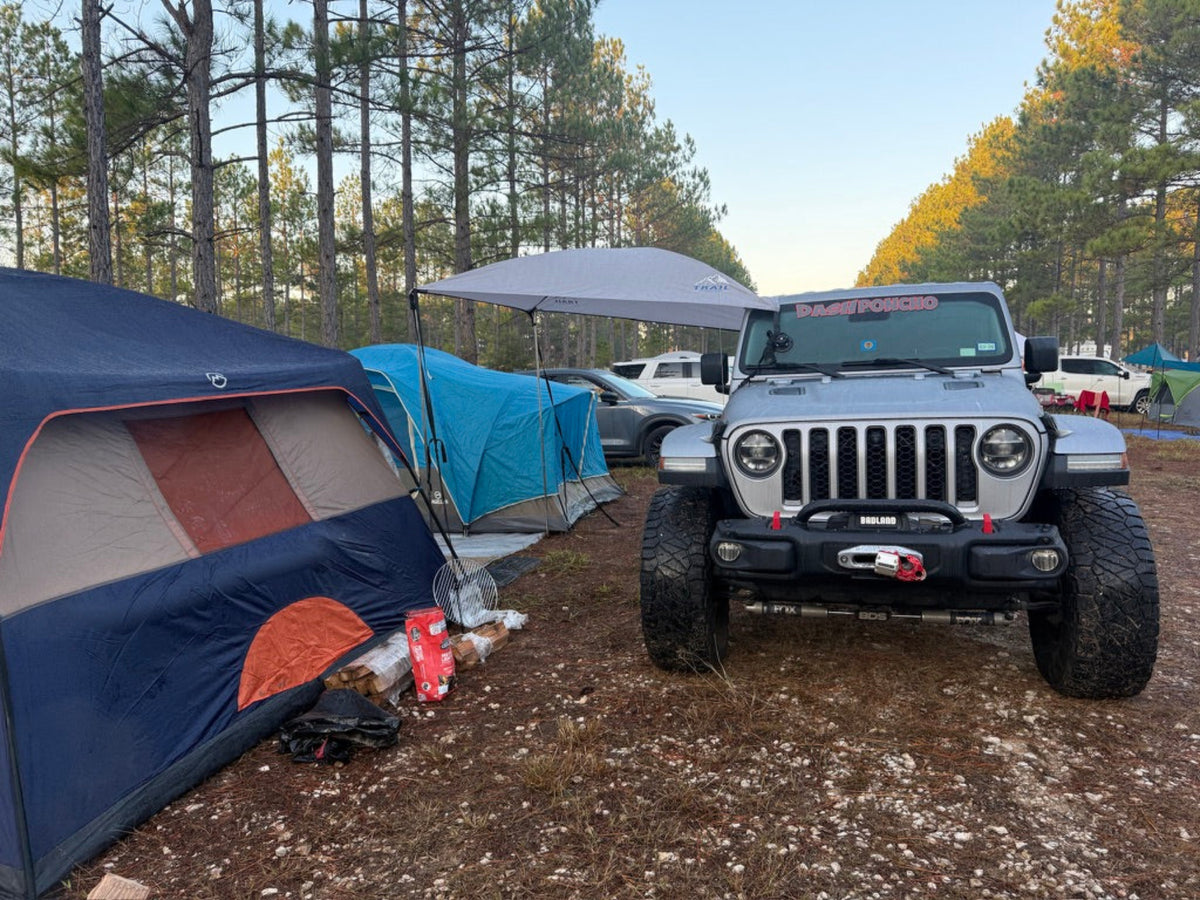 A silver Jeep Wrangler parked at a forest camping site with a blue and orange tent, a blue tarp, and aTrail Topper canopy awning  in the background.