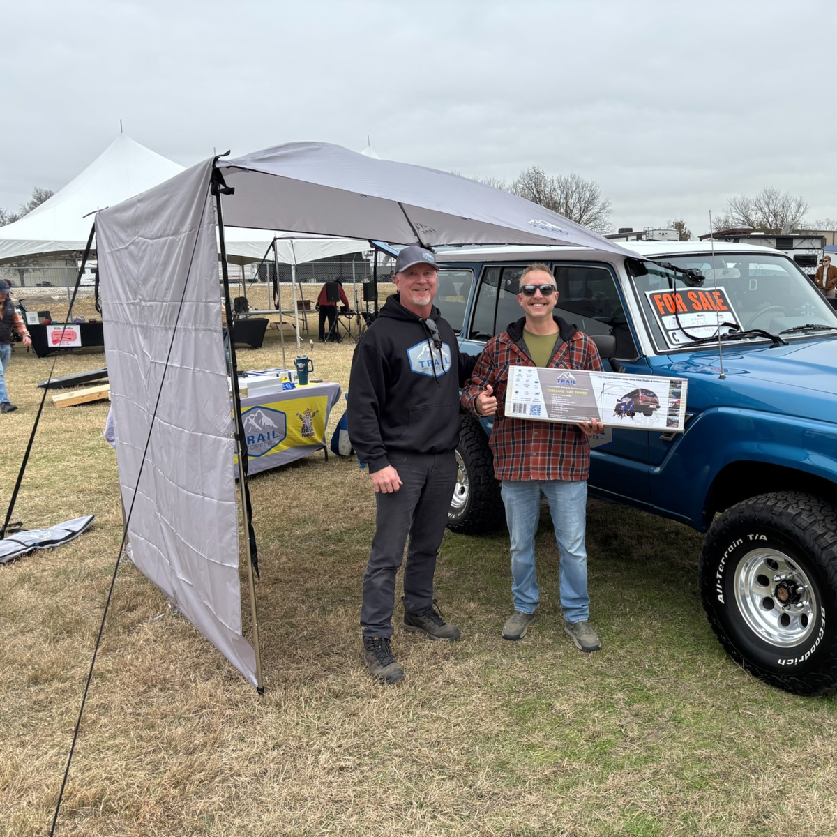 Two men standing under an Overland Trail topper and side wasll with a blue truck in the background