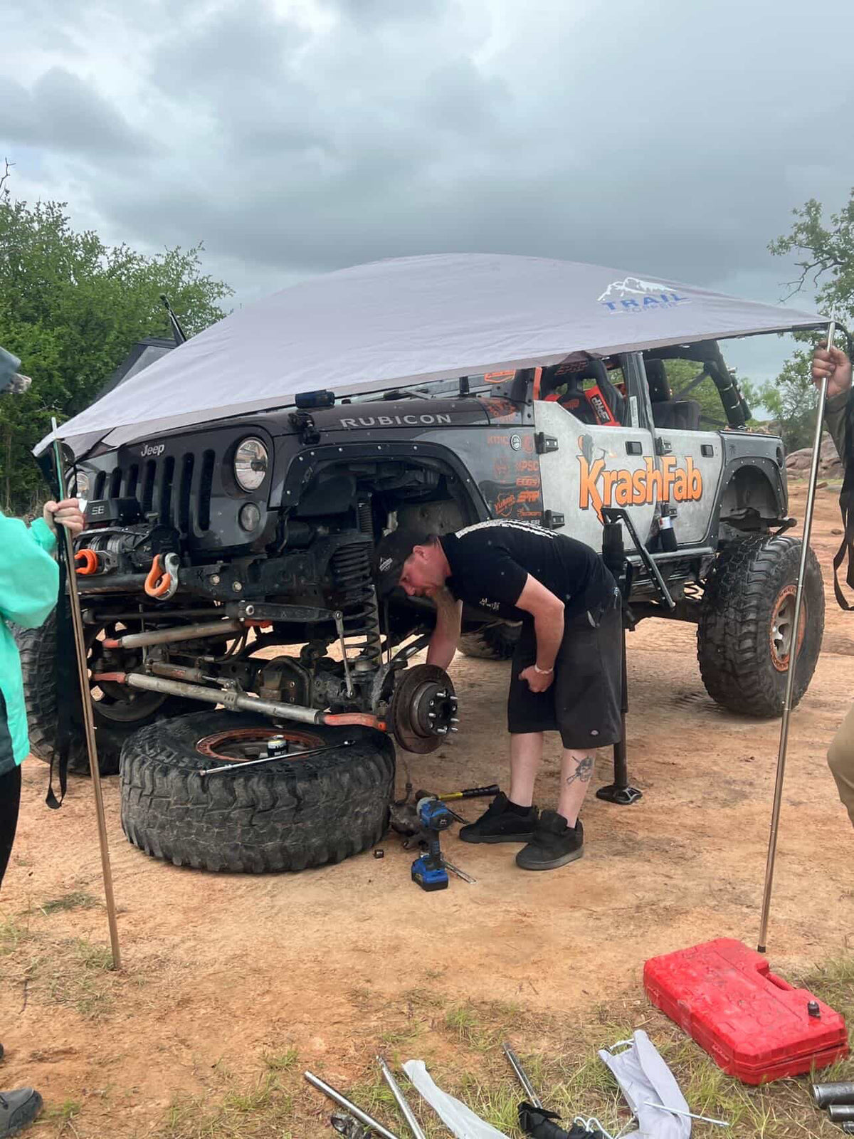 A man in a black t-shirt and shorts works underneath a lifted Jeep with its wheel removed, under a Trail Topper awning on a dirt ground with a cloudy sky in the background.